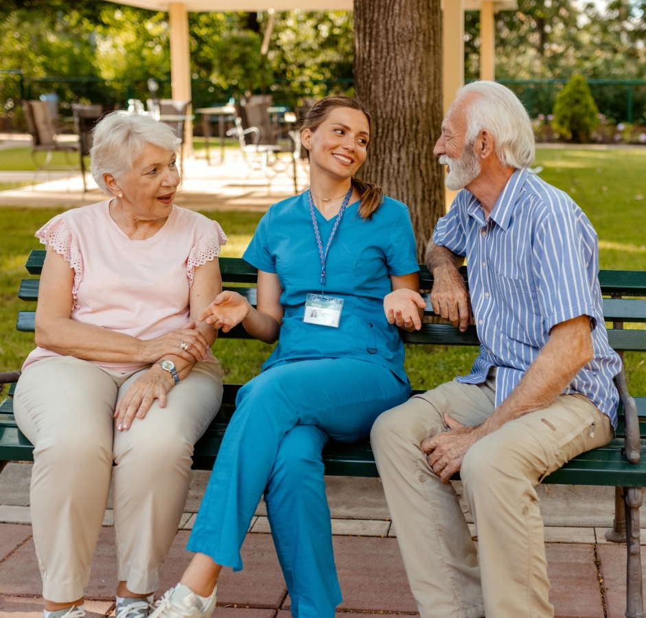 Caregiver talking with elderly couple on bench.