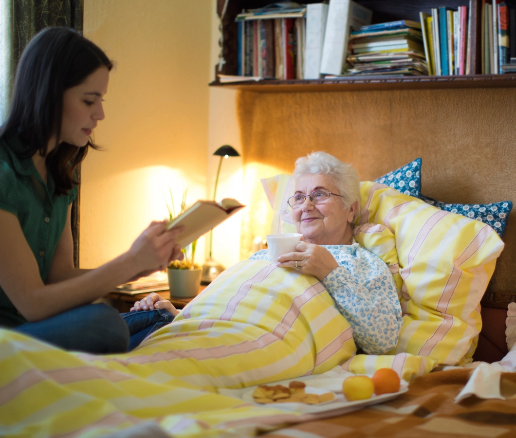 Young woman reading to elderly woman in bed.